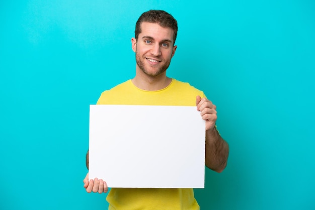 Jeune homme caucasien isolé sur fond bleu tenant une pancarte vide avec une expression heureuse