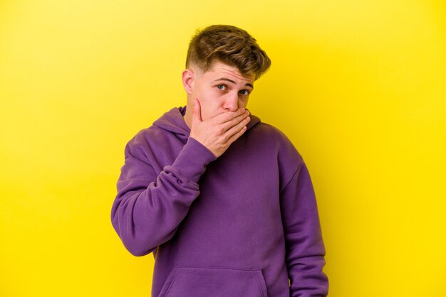 Jeune homme caucasien isolé sur fond jaune couvrant la bouche avec les mains l'air inquiet.