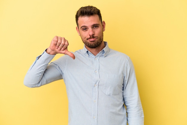 Jeune homme caucasien isolé sur fond jaune montrant un geste d'aversion, les pouces vers le bas. Notion de désaccord.