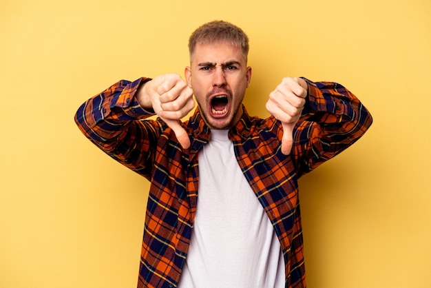 Jeune homme caucasien isolé sur fond jaune montrant le pouce vers le bas et exprimant son aversion.