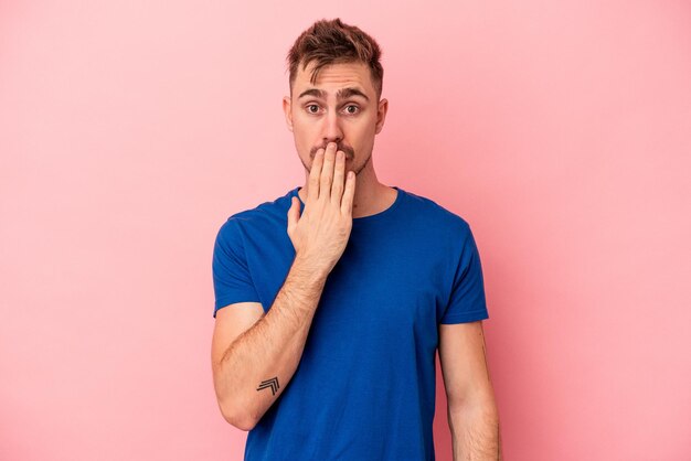 Jeune homme caucasien isolé sur fond rose choqué, couvrant la bouche avec les mains, soucieux de découvrir quelque chose de nouveau.