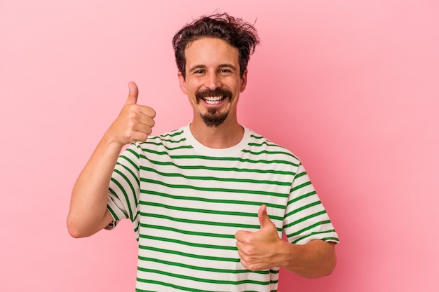 Jeune homme caucasien isolé sur fond rose levant les deux pouces vers le haut, souriant et confiant.