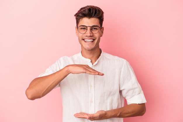 Jeune homme caucasien isolé sur fond rose tenant quelque chose avec les deux mains, présentation du produit.