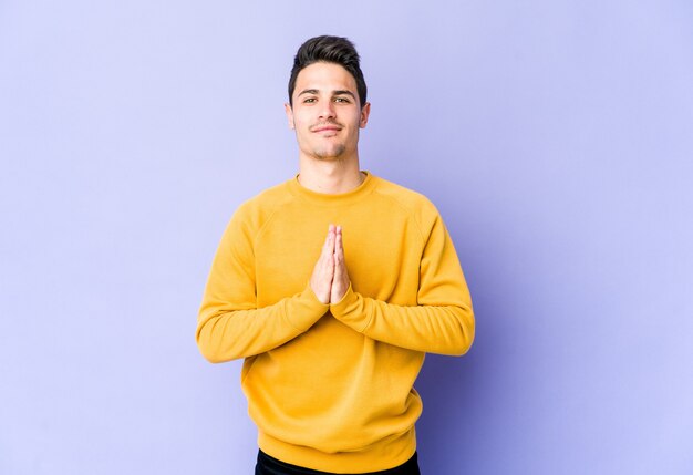 Jeune homme caucasien isolé sur fond violet priant, montrant la dévotion, personne religieuse à la recherche d'inspiration divine.