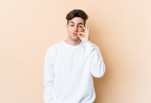 Jeune homme caucasien isolé sur un mur beige avec les doigts sur les lèvres en gardant un secret.