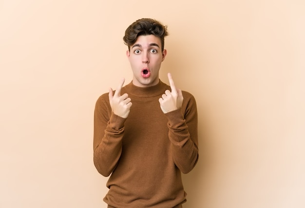 Jeune homme caucasien isolé sur un mur beige pointant vers le haut avec la bouche ouverte.