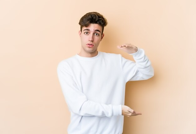 Jeune homme caucasien isolé sur un mur beige tenant quelque chose à deux mains, présentation du produit.