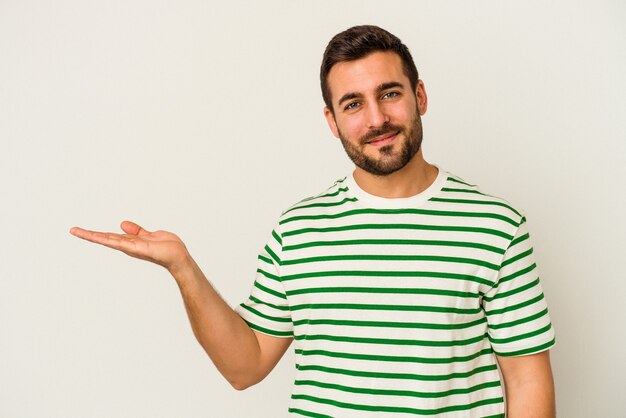 Jeune homme caucasien isolé sur un mur blanc montrant un espace de copie sur une paume et tenant une autre main sur la taille.