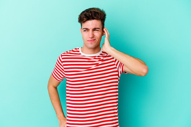 Jeune homme caucasien isolé sur un mur bleu couvrant les oreilles avec les doigts, stressé et désespéré par un bruit ambiant.