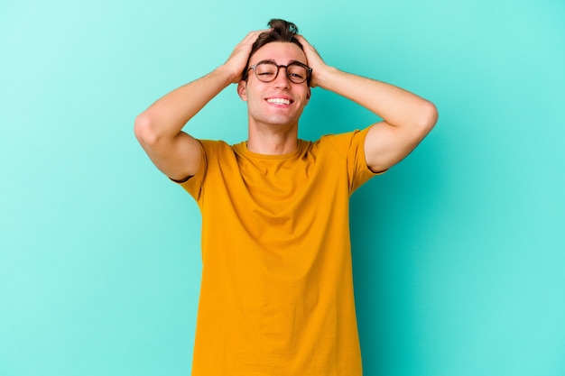 Jeune homme caucasien isolé sur un mur bleu rit joyeusement en gardant les mains sur la tête. Concept de bonheur.