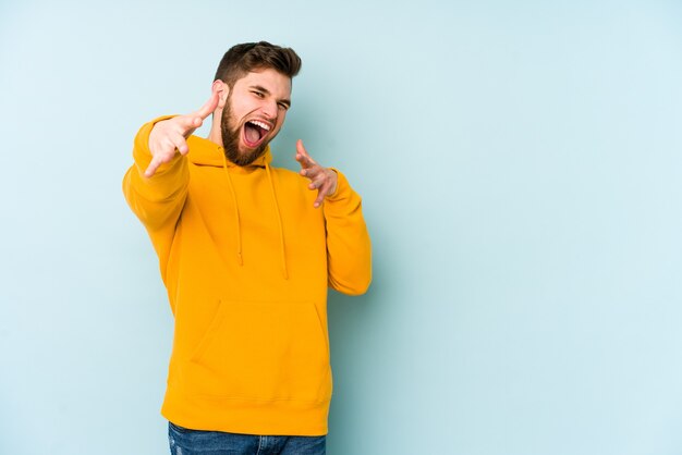 Jeune homme caucasien isolé sur un mur bleu se sent confiant en donnant un câlin à la caméra.