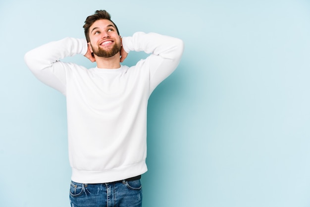 Jeune homme caucasien isolé sur un mur bleu se sentant confiant, avec les mains derrière la tête.