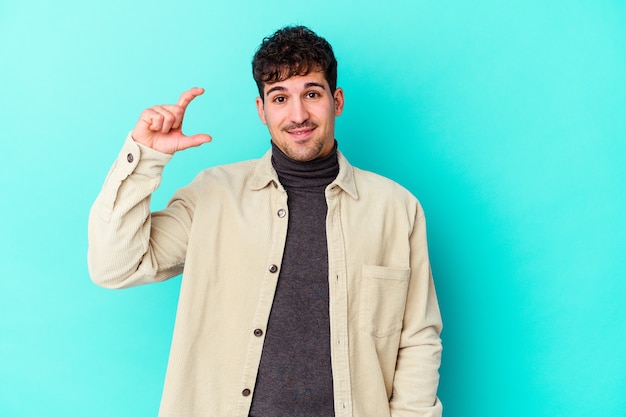 Jeune homme caucasien isolé sur un mur bleu tenant quelque chose de peu avec l'index, souriant et confiant.