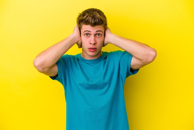 Jeune homme caucasien isolé sur un mur jaune couvrant les oreilles avec les mains en essayant de ne pas entendre un son trop fort.