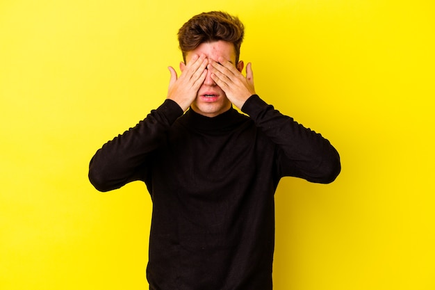 Jeune homme caucasien isolé sur un mur jaune peur couvrant les yeux avec les mains.