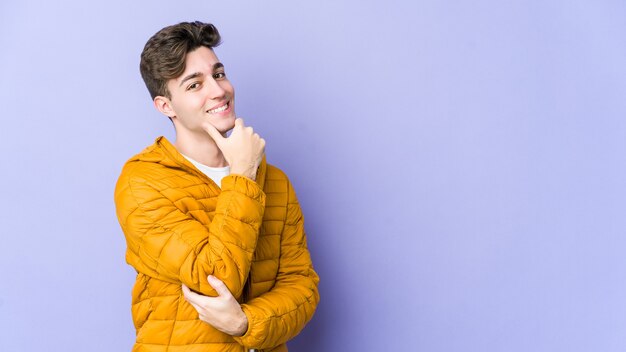 Jeune homme caucasien isolé sur mur violet souriant heureux et confiant, touchant le menton avec la main.