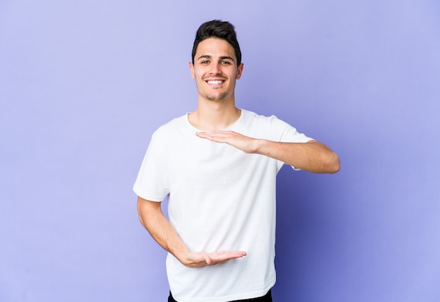 Jeune homme caucasien isolé sur un mur violet tenant quelque chose à deux mains, présentation du produit.