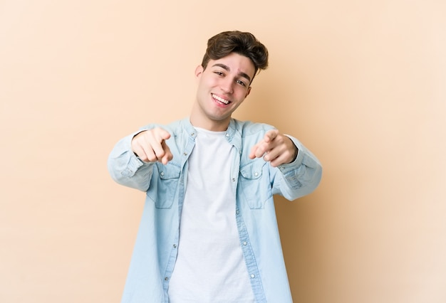 Jeune homme caucasien isolé sur des sourires joyeux mur beige pointant vers l'avant.