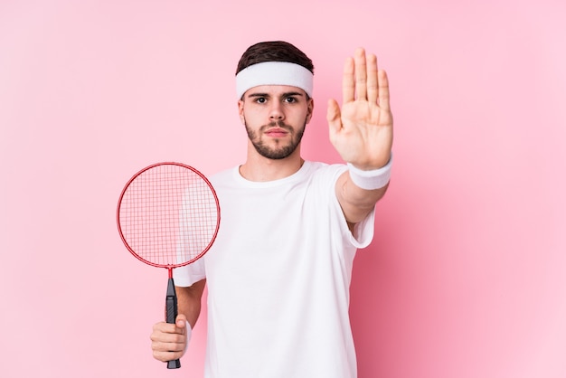 Jeune homme caucasien jouant au badminton debout isolé avec la main tendue montrant le panneau d'arrêt, vous empêchant.