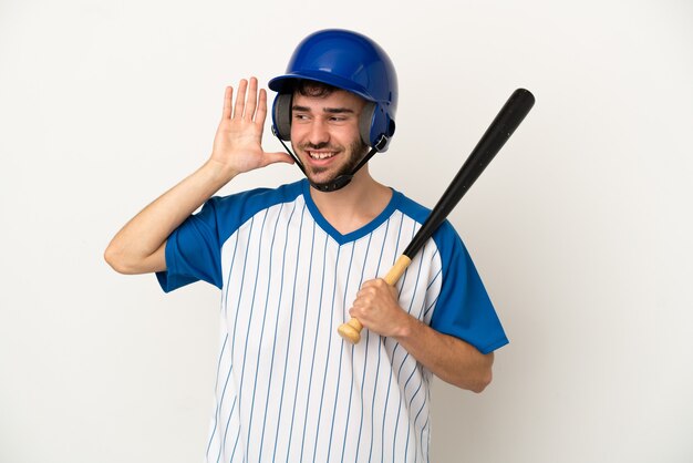 Jeune homme caucasien jouant au baseball isolé sur fond blanc écoutant quelque chose en mettant la main sur l'oreille