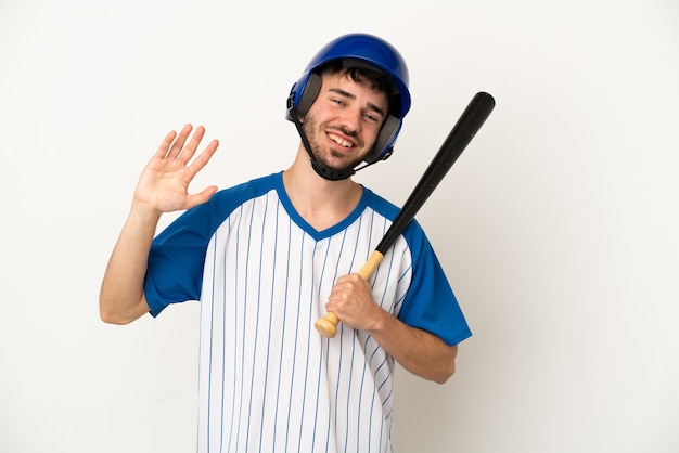 Jeune homme caucasien jouant au baseball isolé sur fond blanc saluant avec la main avec une expression heureuse