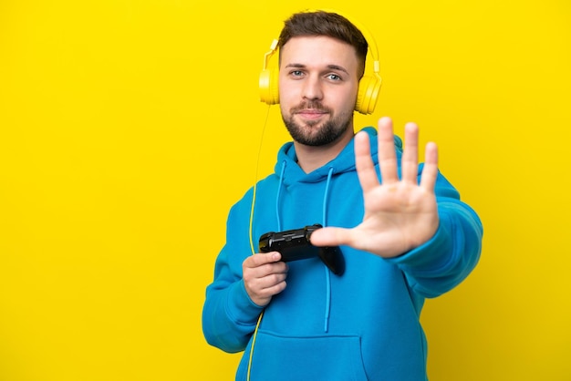 Jeune homme caucasien jouant avec un contrôleur de jeu vidéo isolé sur fond jaune comptant cinq avec les doigts