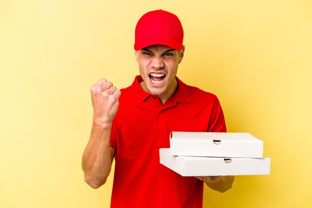 Jeune homme caucasien de livraison tenant des pizzas isolées sur fond jaune montrant le poing à la caméra, expression faciale agressive.
