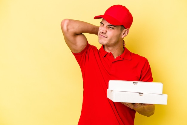 Jeune homme caucasien de livraison tenant des pizzas isolées sur fond jaune touchant l'arrière de la tête, pensant et faisant un choix.