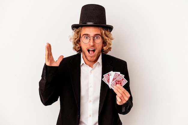 Jeune homme caucasien magicien tenant des cartes magiques isolées sur fond blanc recevant une agréable surprise, excité et levant les mains.
