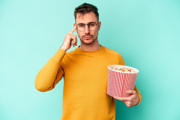 Jeune homme caucasien mangeant du pop-corn isolé sur fond bleu pointant le temple avec le doigt, pensant, concentré sur une tâche.