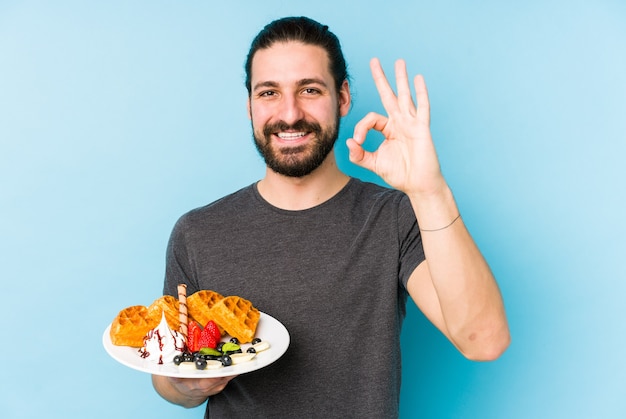 Jeune homme caucasien, manger un dessert gaufré isolé joyeux et confiant montrant le geste ok.