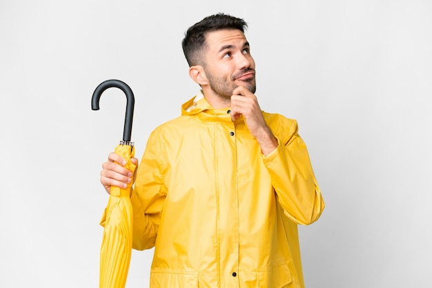 Jeune homme caucasien avec manteau imperméable et parapluie sur fond blanc isolé ayant des doutes