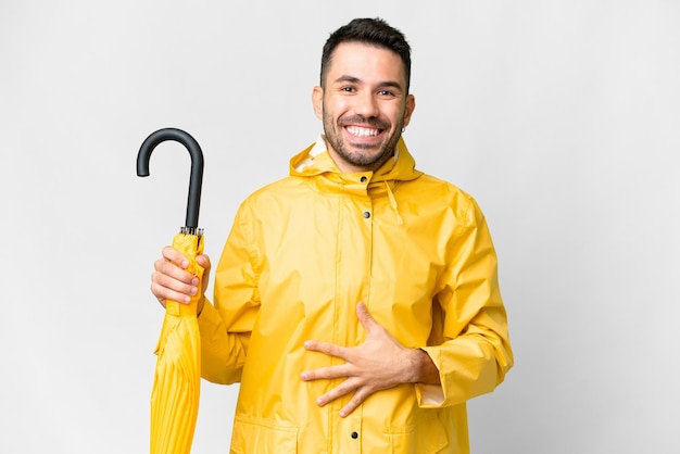 Jeune homme caucasien avec manteau imperméable et parapluie sur fond blanc isolé souriant beaucoup