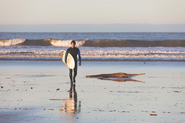 Photo jeune homme caucasien marchant sur la plage avec une planche de surf.