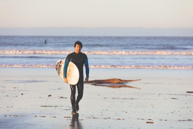 Photo jeune homme caucasien marchant sur la plage avec une planche de surf.