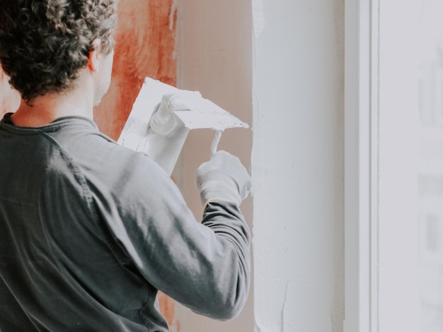 Un jeune homme caucasien met du mastic sur une spatule sur fond de murs préparés avec du mortier rouge