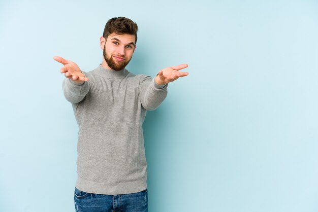 Jeune homme caucasien sur mur bleu fait l'échelle avec les bras, se sent heureux et confiant.