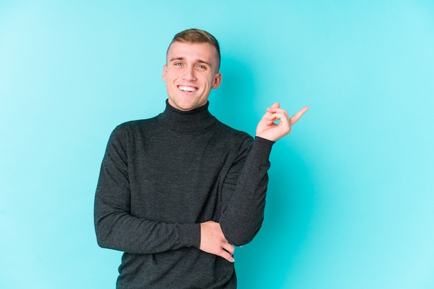 Jeune homme caucasien sur un mur bleu souriant pointant joyeusement avec l'index loin.