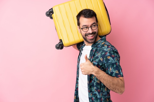 Jeune homme caucasien sur mur isolé en vacances avec valise de voyage et avec le pouce vers le haut