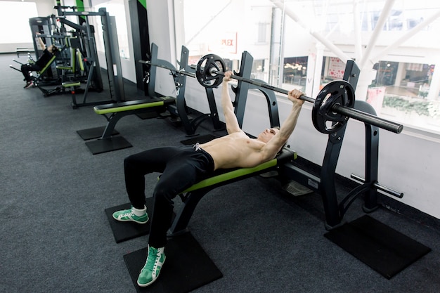 Jeune homme caucasien musclé, soulevant des haltères sur le banc dans la salle de gym.