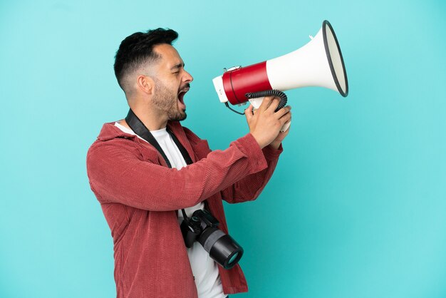 Jeune homme caucasien de photographe d'isolement sur le fond bleu criant par un mégaphone