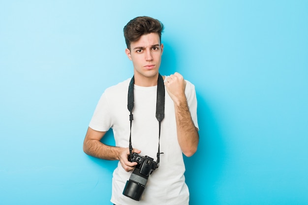 Jeune homme caucasien photographe tenant un appareil photo montrant le poing à la caméra, une expression faciale agressive.