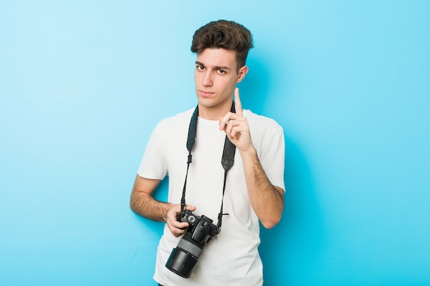 Jeune homme caucasien photographe tenant une caméra montrant le numéro un avec le doigt.