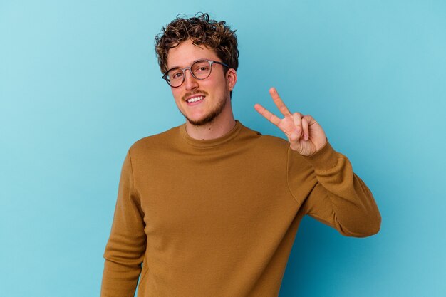 Jeune homme caucasien portant des lunettes isolé sur fond bleu joyeux et insouciant montrant un symbole de paix avec les doigts.