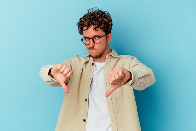 Jeune homme caucasien portant des lunettes isolé sur un mur bleu montrant le pouce vers le bas