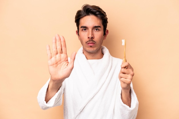 Jeune homme caucasien portant un peignoir tenant une brosse à dents isolé sur fond beige debout avec la main tendue montrant le panneau d'arrêt, vous empêchant.