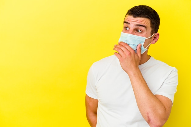 Jeune Homme Caucasien Portant Une Protection Contre Le Coronavirus Sur Jaune Réfléchi à La Recherche D'un Espace De Copie Couvrant La Bouche Avec La Main.