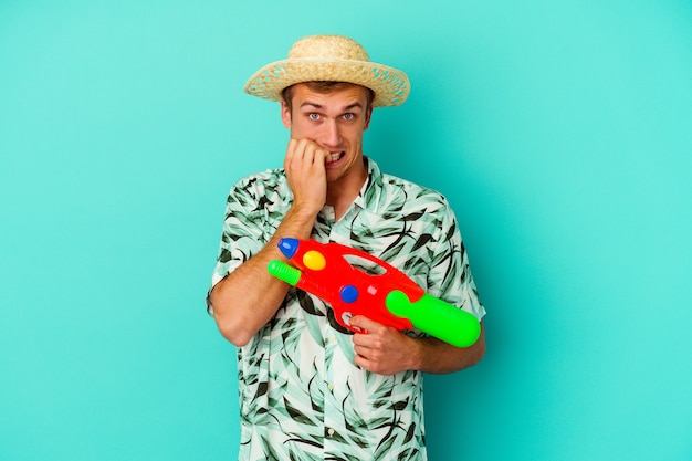 Photo jeune homme caucasien portant des vêtements d'été et tenant un pistolet à eau isolé sur un mur blanc se mordant les ongles, nerveux et très anxieux.