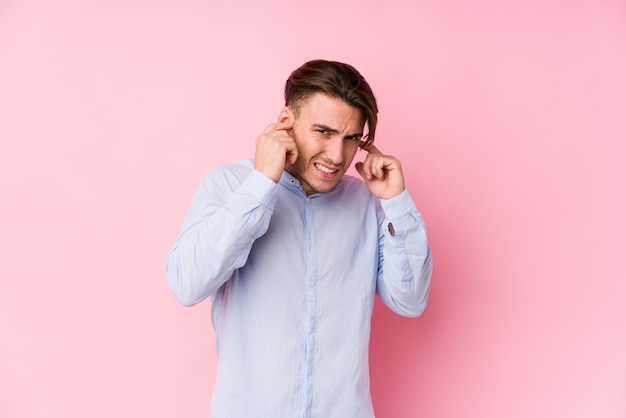 Jeune homme caucasien posant dans un mur rose isolé couvrant les oreilles avec les mains.