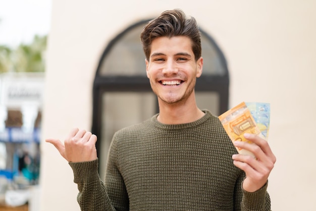 Jeune homme caucasien prenant beaucoup d'argent à l'extérieur pointant vers le côté pour présenter un produit
